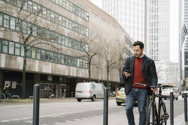 Businessman with bicycle using smart phone while walking against buildings in city, Frankfurt, Germany - AHSF02368