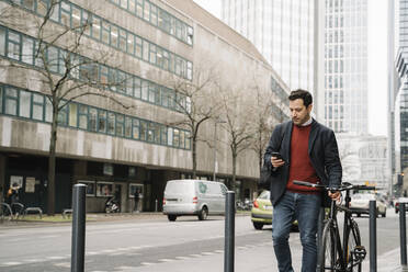 Geschäftsmann mit Fahrrad, der sein Smartphone benutzt, während er gegen Gebäude in der Stadt läuft, Frankfurt, Deutschland - AHSF02368