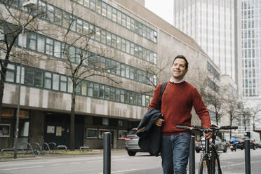 Lächelnder Geschäftsmann, der wegschaut, während er mit dem Fahrrad an der Straße in Frankfurt, Deutschland, spazieren geht - AHSF02369