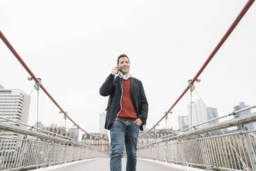 Lächelnder Geschäftsmann, der mit seinem Smartphone spricht, während er auf einer Brücke gegen den Himmel in Frankfurt, Deutschland, läuft - AHSF02371