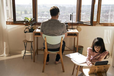 Mädchen macht Hausaufgaben, während ihr Vater im Hintergrund am Laptop arbeitet - ERRF03525