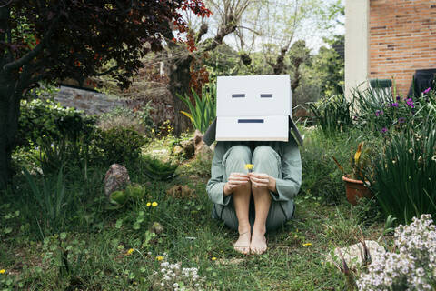 Young woman with cardboard box on her head sitting barefoot in garden stock photo