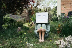 Young woman with cardboard box on her head sitting barefoot in garden - AFVF06131