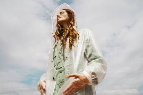 Redheaded young woman with eyes closed wearing transparent rain coat standing against sky stock photo