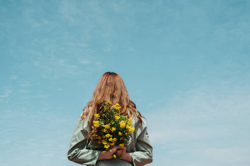 Back view of redheaded young woman against sky holding bunch of flowers behind her back - AFVF06146