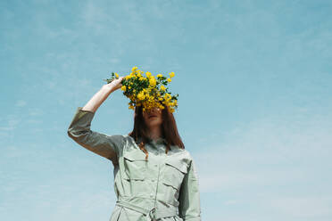 Redheaded young woman against sky covering eyes with bunch of yellow flowers - AFVF06147