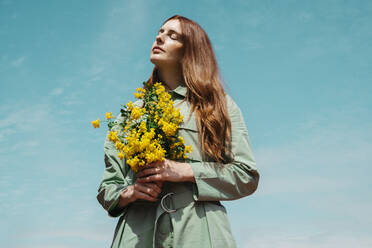 Portrait of redheaded young woman with eyes closed standing against sky holding bunch of yellow flowers - AFVF06148
