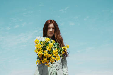Porträt einer rothaarigen jungen Frau mit einem gelben Blumenstrauß vor dem Himmel - AFVF06149