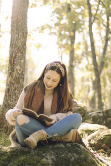 Junge Frau liest ein Buch im Sitzen und umgeben von einem Wald - CAVF80461