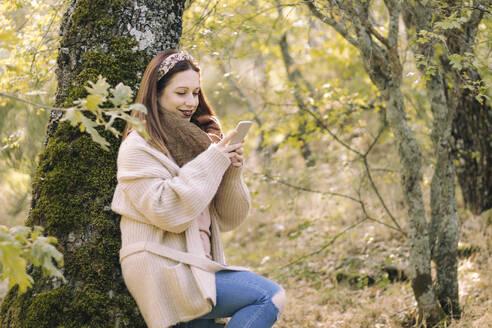 Lächelnde junge Frau, die an einem Baum lehnt und ihr Mobiltelefon benutzt - CAVF80463