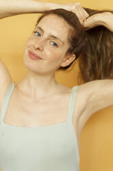 Porträt einer lächelnden rothaarigen Frau, die ihre Haare vor einem gelben Hintergrund zusammenbindet - AFVF06163