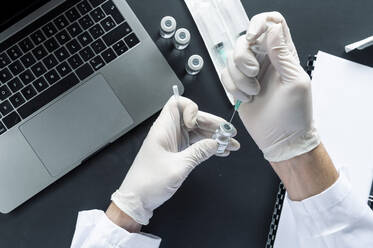 Ausgeschnittenes Bild der Hand eines Arztes, der eine Spritze in ein Fläschchen über einem Laptop am Labortisch injiziert - JCMF00643