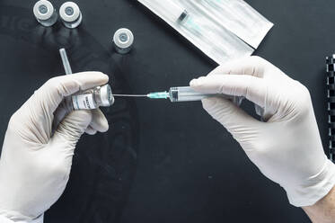 Cropped image of doctor's hand injecting syringe in vaccination vial over laptop at laboratory desk - JCMF00644