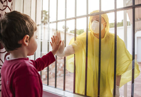 Boy touching mother's hand wearing protective clothing behind windowpane - LJF01513