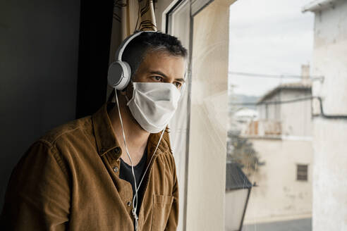 Mann mit Schutzmaske und weißen Kopfhörern schaut aus dem Fenster - RCPF00234