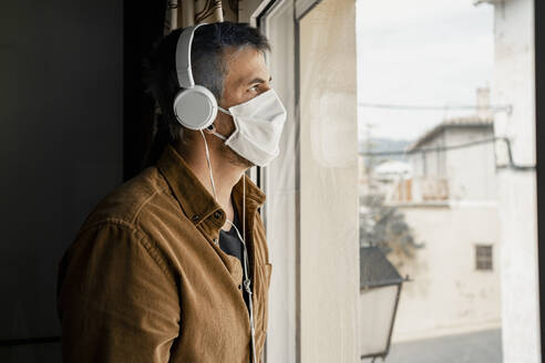 Man wearing protective mask and white headphones looking out of the window - RCPF00235