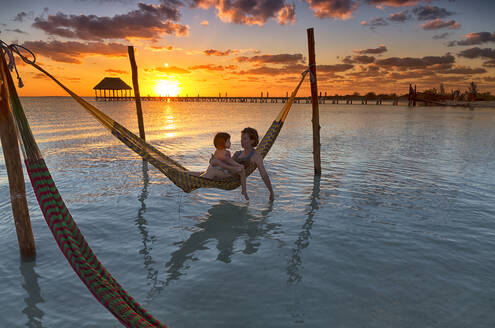 Mutter und Tochter entspannen sich in einer Hängematte am Strand bei Sonnenuntergang auf der Insel Holbox, Cancun, Mexiko - DSGF02015