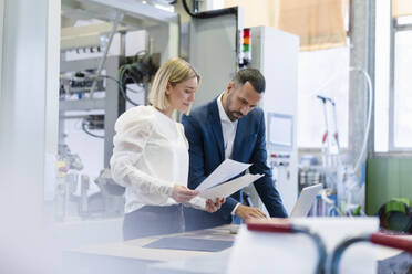 Businessman and young woman with papers and laptop talking in a factory - DIGF09888