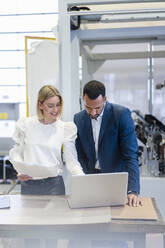 Geschäftsmann und junge Frau mit Papieren und Laptop im Gespräch in einer Fabrik - DIGF09889