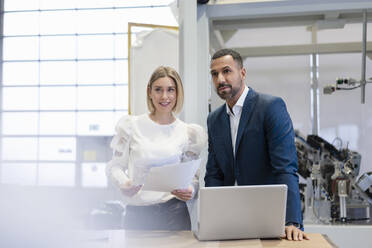 Geschäftsmann und junge Frau mit Papieren und Laptop in einer Fabrik - DIGF09890