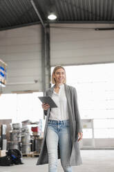 Smiling young woman holding tablet walking in a factory - DIGF09910