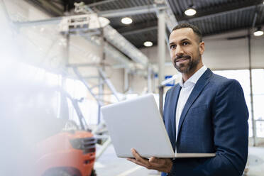 Smiling businessman holding laptop in a factory - DIGF09930