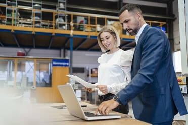 Businessman and young woman with papers and laptop in a factory - DIGF09934