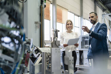 Geschäftsmann und junge Frau mit Papieren im Gespräch an einer Maschine in einer Fabrik - DIGF09940