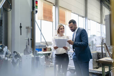 Geschäftsmann und junge Frau mit Papieren im Gespräch an einer Maschine in einer Fabrik - DIGF09942
