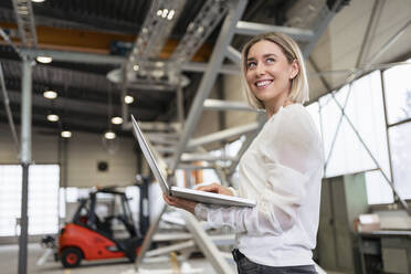 Lächelnde junge Frau mit Laptop in einer Fabrik - DIGF09969