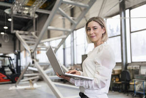 Porträt einer lächelnden jungen Frau mit Laptop in einer Fabrik - DIGF09970