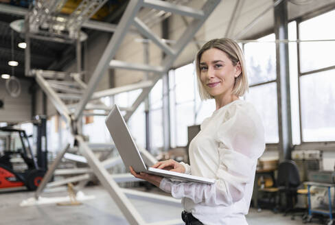 Portrait of smiling young woman using laptop in a factory - DIGF09970