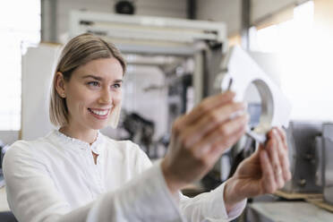 Smiling young woman holding workpiece in a factory - DIGF09986