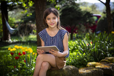 Porträt eines lächelnden Mädchens, das mit einem digitalen Tablet im Garten sitzt - LVF08860