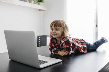 Kleines Mädchen liegt auf dem Schreibtisch und schaut auf den Laptop - JRFF04387