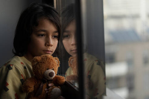 Boy with his teddy bear looking out of window at home - VABF02827