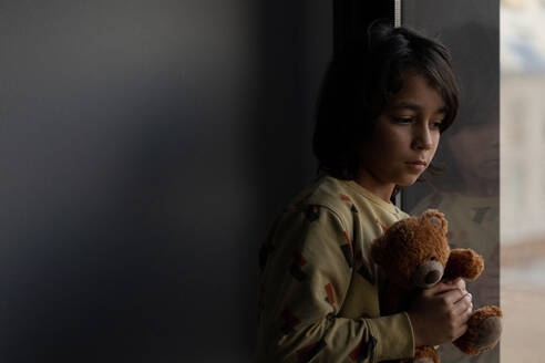 Boy with his teddy bear looking out of window at home - VABF02828