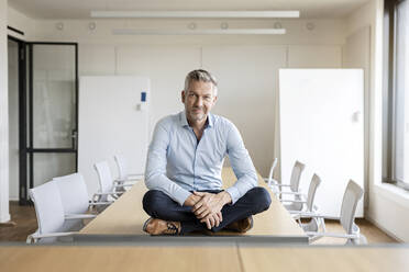 Porträt eines selbstbewussten reifen Geschäftsmannes, der auf einem Tisch in einem Konferenzraum sitzt - PESF01993