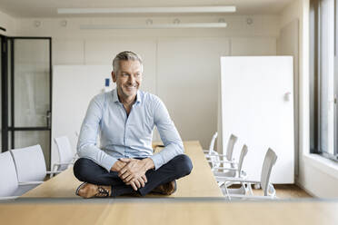 Portrait of smiling mature businessman sitting on table in conference room - PESF01994