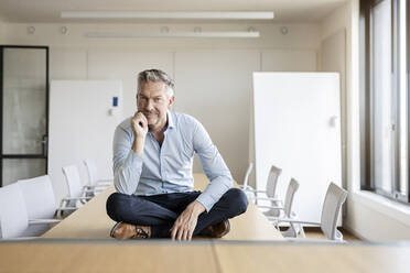 Porträt eines selbstbewussten reifen Geschäftsmannes, der auf einem Tisch in einem Konferenzraum sitzt - PESF01995