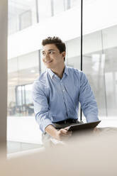 Porträt eines lächelnden jungen Geschäftsmannes mit Tablet am Fenster im Büro - PESF02034