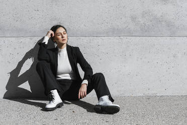 Young woman wearing black suit sitting on floor in front of concrete wall - TCEF00517