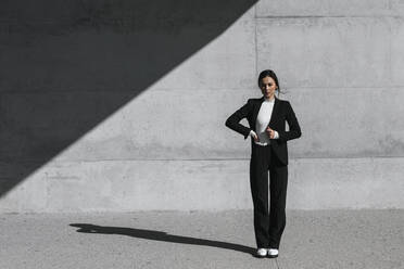Junge Frau in schwarzem Anzug steht vor einer Betonwand - TCEF00526