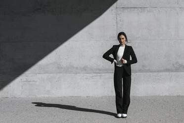 Young woman wearing black suit standing in front of concrete wall - TCEF00526