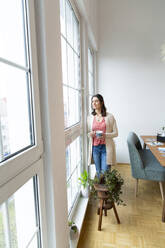 Woman standing at the window at home looking out - FKF03756