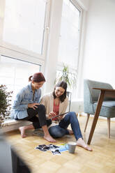 Happy mother and daughter sitting on the floor at home looking at photographs - FKF03760