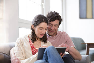 Couple sitting on couch at home using tablet - FKF03766