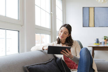 Woman sitting on couch at home using tablet - FKF03770