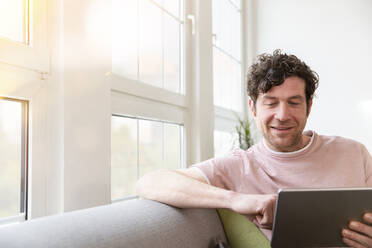 Man sitting on couch at home using tablet - FKF03771