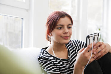 Teenager-Mädchen mit Smartphone und Kopfhörern sitzt zu Hause auf der Couch - FKF03774